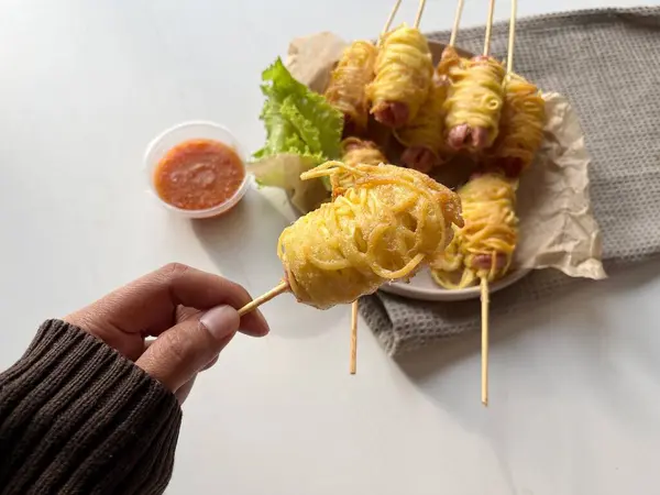 Sosis mie gulung 'u tutan bir el. Tipik Endonezya atıştırmalıkları. Rulo erişteli sosis sosislerden yapılır. Sosisler kıvrıldıktan sonra kuru eriştenin içine sarılır ve çırpılmış yumurta ile kızartılır..