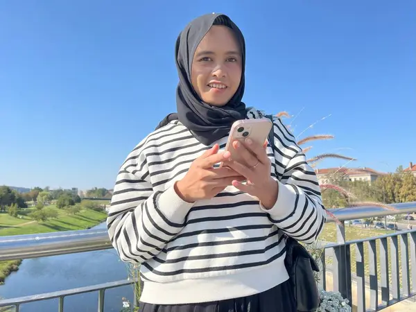 Çizgili kapüşonlu genç Asyalı bir kadın köprüde kameraya bakıyor, akıllı telefonunu canlı mavi gökyüzünün arka planına karşı kullanarak gülümsüyor.