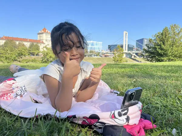 Asyalı küçük bir kız çimlerin üzerinde telefon kullanıyor ve el hareketleriyle video görüşmesi yapıyor.