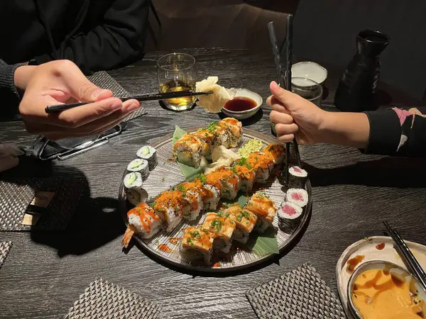 Suşi yemeğinin tadını çıkaran iki kişi: Büyük bir tabaktan suşi parçaları toplamak için çubuklar kullanıyorlar, bu tabakta çeşitli dürüm ve nigiri çeşitleri yer alıyor..