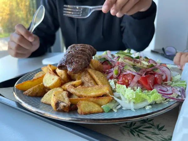 Plastik çatal tutan tanınmamış bir adam. Restoranda servis edilen kebap, patates kızartması ve beyaz bir tabakta salata ile birlikte.