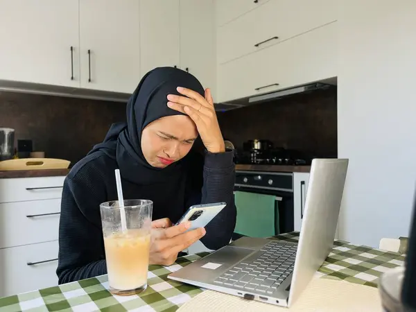 Ev mutfağında tesettürlü Asyalı Müslüman bir kadın telefon ve dizüstü bilgisayar kullanıyor. Yüzü asık ve telefona bakıyor. Laptopun yanında buzlu kahve.