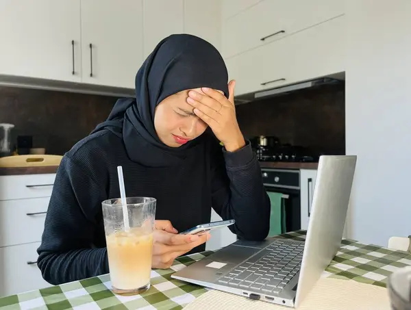 Ev mutfağında tesettürlü Asyalı Müslüman bir kadın telefon ve dizüstü bilgisayar kullanıyor. Yüzü asık ve telefona bakıyor. Laptopun yanında buzlu kahve.