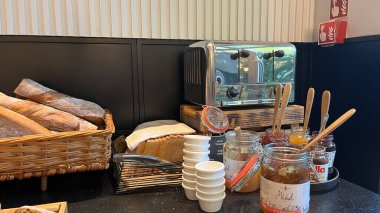 Varşova, Polonya. 02 Kasım 2024 'te çeşitli ekmek seçenekleri, yayılma ve tost makinesi olan bir kahvaltı büfesi. Farklı ekmek türleri, reçel kavanozları ve bal dolu sepetler var.