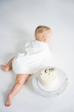 Beyaz elbiseli bir bebek beyaz zeminde bir tepesiyle küçük bir doğum günü pastasının yanında emekliyor, ilk doğum gününü kutluyor.
