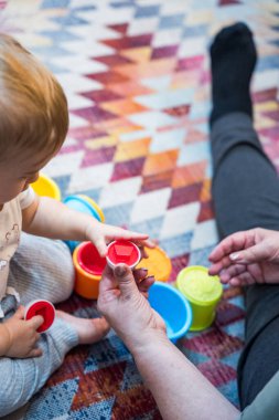Bebek ve yetişkin evdeki desenli halı üzerinde renkli bardaklarla oynuyorlar, erken çocukluk gelişimini teşvik ediyorlar ve öğreniyorlar.