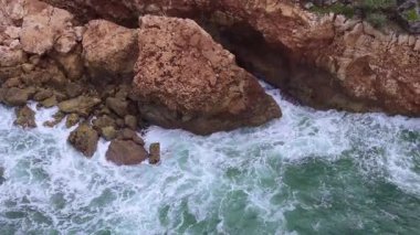 Static aerial shot captures foamy turquoise waves churning around and crashing against a cluster of large, rugged coastal rocks and cliff formations. High quality 4k footage
