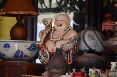 A smiling, jovial Buddha statue with a large belly and beaded necklace sits amongst various antique and decorative items in a cluttered display. High quality photo
