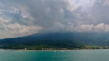 Geniş bir hava manzarası, bulutlu bir gökyüzünün altındaki büyük, puslu bir dağın eteğinde yuva yapmış bir köy ile canlı yeşil tropikal bir sahil şeridi. Yüksek kaliteli illüstrasyon