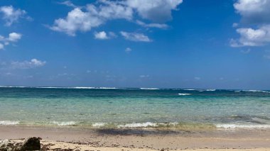 Sakin, berrak turkuaz bir okyanus suyuyla geniş, sakin bir sahil manzarası dağınık bulutlu, parlak mavi bir gökyüzünün altında yavaşça kumlu bir sahili kaplıyor. Yüksek kalite fotoğraf