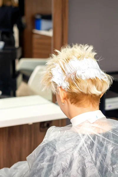 Coloring white hair with hair dye of the young caucasian blonde woman sitting at a hair salon, close up
