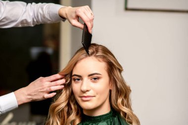 Kuaför, uzun beyaz kadın saçlarını tarıyor kameraya bakıyor ve güzellik salonunda gülümsüyor.