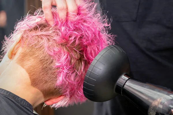 Drying short pink bob hairstyle of a young caucasian woman with a black hair dryer with the brush by hands of a male hairdresser in a hair salon, close up