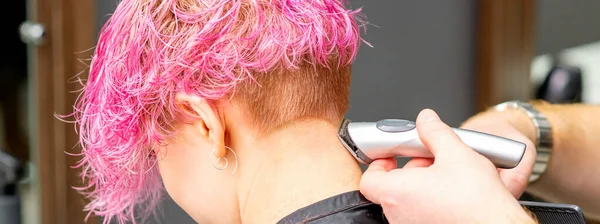 在美发厅里 一名年轻的高加索女人用电动剃须刀剃短粉色的头发 他的男性理发师用电动剃须刀刮她的脖子 — 图库照片