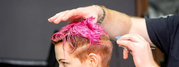 理发店里的一位男性理发师把年轻的高加索女人染成粉色的短而湿的头发剪下来 — 图库照片