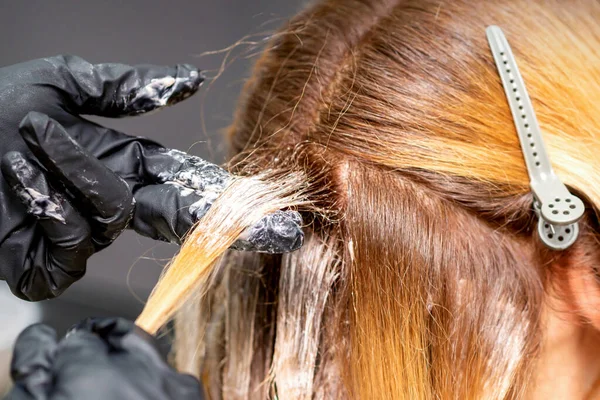 理发师的手正在用戴着手套的手把染发剂涂在一位红头发的年轻女子的红头发上 她正在美发厅里闭着眼睛 — 图库照片