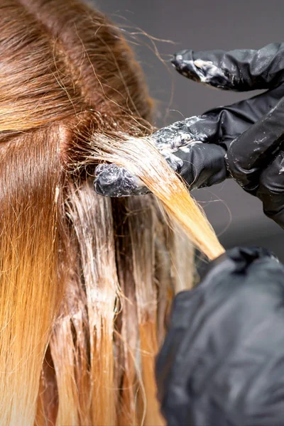 理发师的手正在用戴着手套的手把染发剂涂在一位红头发的年轻女子的红头发上 她正在美发厅里闭着眼睛 — 图库照片