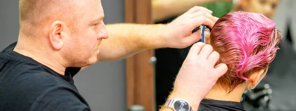 理发店里的一位男性理发师把年轻的高加索女人染成粉色的短而湿的头发剪下来 — 图库照片