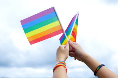 Asyalı bir kadın gökyüzü arka planında bir gökkuşağı bayrağı tutuyor, üzerinde PRIDE mesajı ve gökkuşağı bilekliği yazılı siyah bir bileklik takıyor. lgbt gururunun sembolü                                 