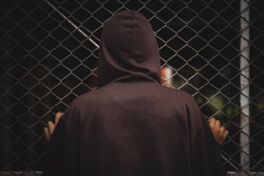 Kahverengi kapüşonlu genç bir erkek çelik tel örgüye bakıyordu. İki el de çitlere tutunup arkasını gösteriyor..                               