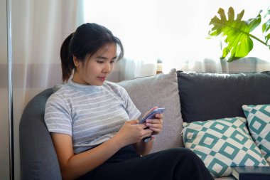 Asyalı bir kadın oturma odasında kanepede oturmuş arkadaşlarıyla cep telefonuyla sohbet ediyor. Boş zaman aktivitesi kavramı, dinlenme, tatil. Yumuşak ve seçici odak.                               