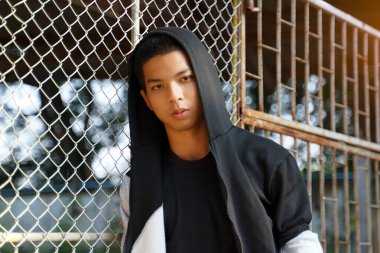 young man wearing a hoodie sits leaning against a wire mesh fence. Sitting blankly, separated from friends. Concept of teenagers, having problems, thinking, alone.                                clipart