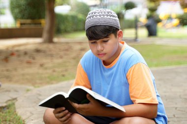 Genç Müslüman bir çocuk mavi bir tişört ve gri bir taksi şapkası giyiyor. Parkta kitap okurken oturup poz vermek..                               