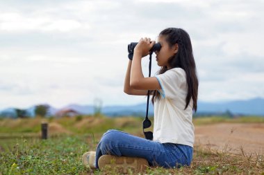 Asyalı kız sahada doğayı keşfetmek için dürbün kullanıyor. Sınıf dışında öğrenme kavramı doğa çalışması, keşif.                                