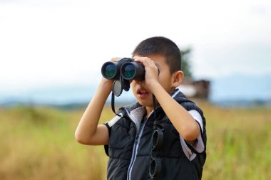 Asyalı çocuk sahada doğayı keşfetmek için dürbün kullanıyor. Sınıf dışında öğrenme kavramı doğa çalışması, keşif.                                