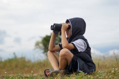 Asyalı çocuk sahada doğayı keşfetmek için dürbün kullanıyor. Sınıf dışında öğrenme kavramı doğa çalışması, keşif.                                
