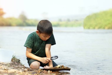 Asyalı çocuk nehirde bulunan taşları incelemek için büyüteç kullanıyor. Açık hava aktiviteleri, keşif, doğa, öğrenme kavramı.                         