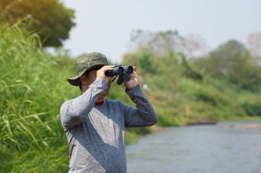Asyalı adam nehir boyunca yerel kuşları gözlemlemek için dürbün kullanıyor. Yumuşak ve seçici odak.                                
