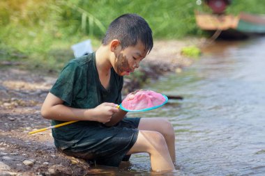 Çamurlu yanaklı Asyalı çocuk nehir kıyısında oturmuş nehirden çıkarılan eleğin içindekileri izliyor.                               