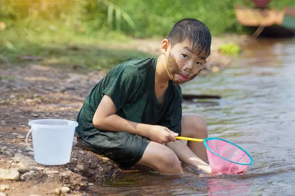 Çamurlu yanaklı Asyalı çocuk nehir kıyısında oturmuş nehirden çıkarılan eleğin içindekileri izliyor..                                