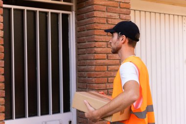 Turuncu yelek giymiş genç ve çekici bir adam şehir ve şehir geçmişinde paket teslimatçısı olarak çalışıyor.