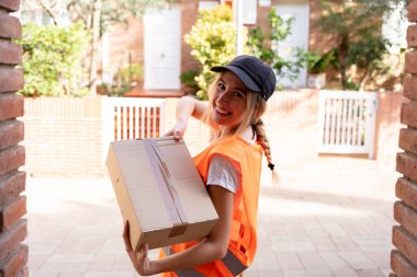 Karton bir kutuyla müşteriye teslim edilecek genç bir teslimatçı kız. Mutlu görünüyor ve gülümsüyor.