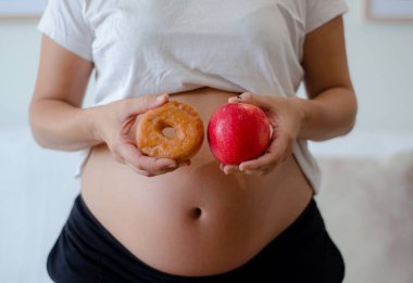 Elinde donut ve elmayla hamile bir kadının karnını kapatıyoruz. Kişisel bakım, doğum öncesi diyet ve sağlıklı alışkanlıklar konsepti. Reklam için kopyalama alanı