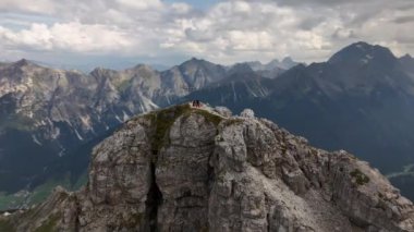 İnsanlar bir dağın tepesinde oturuyorlar. Arkasında dağlar ve vadiler var. Yörüngesel dron videosu. Hoher Burgstall, Yedi Zirve Stubai, Tirol, Avusturya..