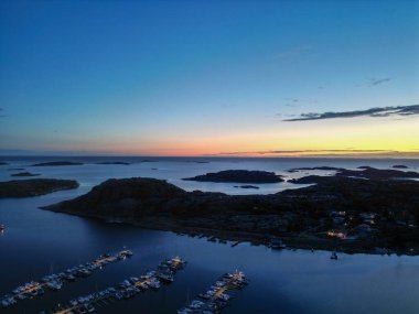 A serene twilight view of a marina with boats docked, surrounded by rocky islands and calm waters. The sky transitions from blue to orange as the sun sets on the horizon. clipart