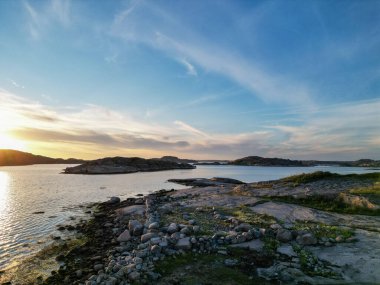 Kayalık kıyılar, sakin sular ve gün batımında renkli bir gökyüzü içeren sakin bir kıyı manzarası. Ön planda dağınık taşlar ve yeşillikler yer alırken, ufukta uzak adalar görülebilir..