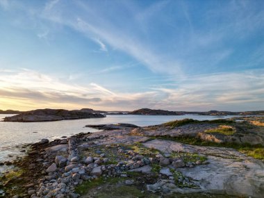Kayalık kıyılar, sakin sular ve gün batımında renkli bir gökyüzü içeren sakin bir kıyı manzarası. Ön planda dağınık taşlar ve yeşillikler yer alırken, ufukta uzak adalar görülebilir..
