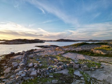 Kayalık kıyılar, sakin sular ve gün batımında renkli bir gökyüzü içeren sakin bir kıyı manzarası. Ön planda dağınık taşlar ve yeşillikler yer alırken, ufukta uzak adalar görülebilir..