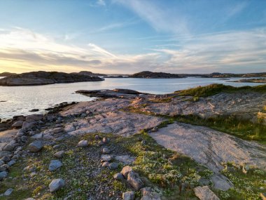 Kayalık kıyılar, sakin sular ve gün batımında renkli bir gökyüzü içeren sakin bir kıyı manzarası. Ön planda dağınık taşlar ve yeşillikler yer alırken, ufukta uzak adalar görülebilir..