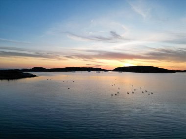 Sakin bir denizin üzerinde sakin bir günbatımı. Birkaç küçük ada ve demir atmış yelkenliler. Gökyüzü sıcacık turuncu ve sarı renklerle boyanmıştır, suyun yüzeyini yansıtır..