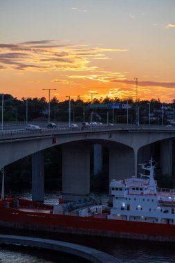 Gün batımı manzarasında araçlı bir köprü ve altından geçen büyük bir gemi var. Gökyüzü sıcak turuncu ve sarıya boyanmıştır, suyu yansıtır. Sahne sakin bir şehir akşam atmosferini yakalıyor..