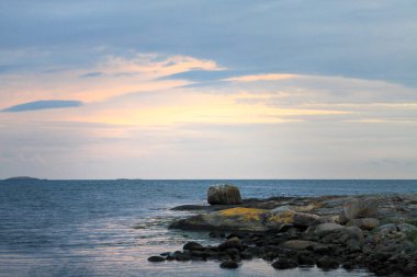 Sakin suların, kayalık kıyıların ve günbatımında pastel gökyüzünün olduğu sakin bir kıyı manzarası. Sahne, doğanın sükunetini yumuşak dalgalar ve dağınık kayalarla yakalıyor..