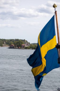 Rüzgarda dalgalanan bir İsveç bayrağı, manzaralı bir göl ve arka planda ağaçlar. Gökyüzü parçalı bulutlu, sakin bir atmosfer yaratıyor..