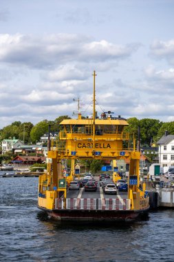 Castella adında sarı bir feribot rıhtıma geliyor, içinde arabalar var. Sahnede yeşil ağaçlar ve arkaplanda parçalı bulutlu bir gökyüzünün altında binalarla çevrili sakin bir su yolu yer alıyor..