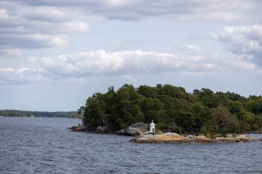 Bereketli yeşil ağaçları ve beyaz deniz feneri olan küçük bir adanın olduğu huzurlu bir sahil manzarası. Sakin sular bulutlu gökyüzünü yansıtıyor ve sakin bir atmosfer yaratıyor..