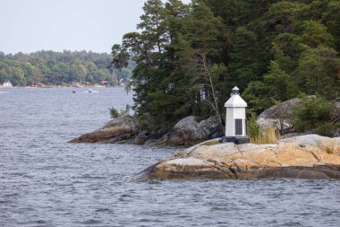 Ağaçlar ve suyla çevrili kayalık bir sahildeki deniz fenerinin manzarası. Deniz feneri siyah tabanlı beyaz, büyük bir kayanın üzerinde duruyor. Arka planda, kayıklar ve yemyeşil bir arazi var..